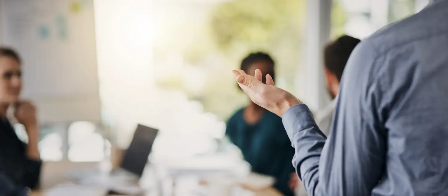 Meetingraum mit verschiedenen Personen, eine vortragende Person wird von hinten gezeigt